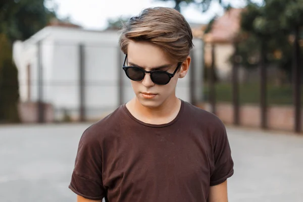 Urban American hipster jeune homme en lunettes de soleil en t-shirt tendance avec coiffure sur un terrain de basket moderne à l'extérieur. Beau mec attrayant en vacances . — Photo