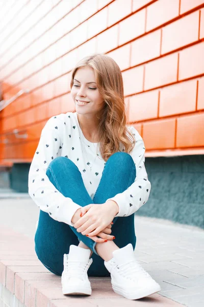 Привлекательная молодая веселая женщина в винтажном свитере в модных джинсах в стильных кроссовках сидит на земле рядом с современным оранжевым зданием на улице. Милая блондинка отдыхает. . — стоковое фото