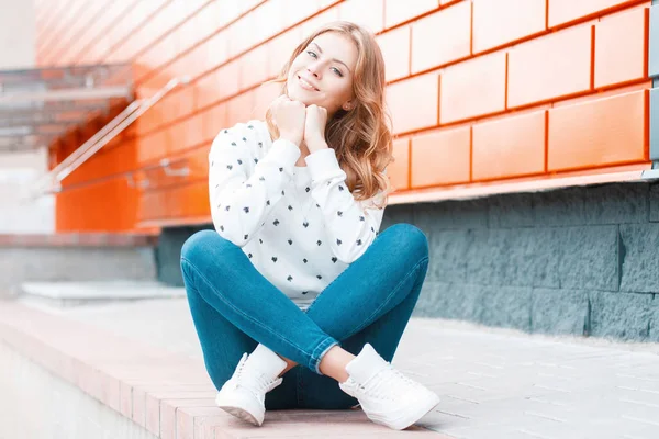 Jovem europeia alegre mulher alegre em um suéter vintage em jeans na moda em tênis senta-se e sonha perto do edifício de tijolo moderno na rua. Menina loira muito feliz goza de um fim de semana . — Fotografia de Stock