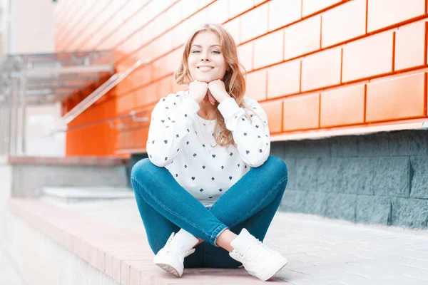 Привлекательная стильная веселая молодая женщина со сладкой улыбкой в винтажных джинсах в кроссовках в свитере сидит на плитке рядом с кирпичным зданием. Позитивная девушка на улице. Весенняя модная молодежная одежда . — стоковое фото