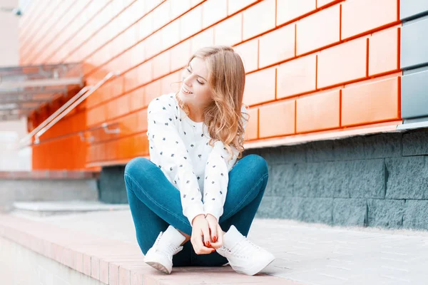 Забавная современная девушка в стильных кроссовках в винтажных джинсах в элегантном свитере сидит на тротуаре рядом с современным черно-оранжевым зданием. Милая симпатичная девушка-модель в отпуске . — стоковое фото