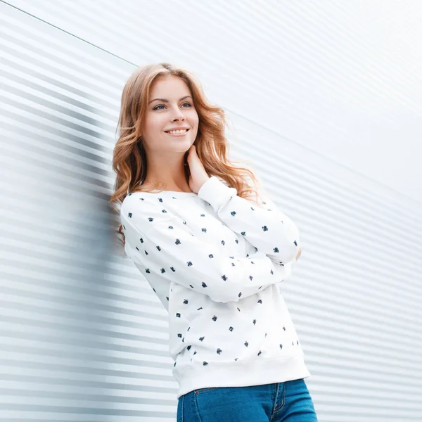 Trendy mavi kot trendy beyaz kazak pozitif mutlu genç kadın açık havada modern beyaz metal duvarın yakınında duruyor. Güzel tatlı kız bir bahar gününde büyük hava sahiptir. — Stok fotoğraf