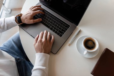 Genç serbest çalışan adam kişisel blogunu internette yönetiyor. Modern bir dizüstü bilgisayar üzerinde çalışan bir iş adamı. Üzerinde kahve, dizüstü bilgisayar, not defteri olan beyaz bir masaüstüne yakın çekim.
