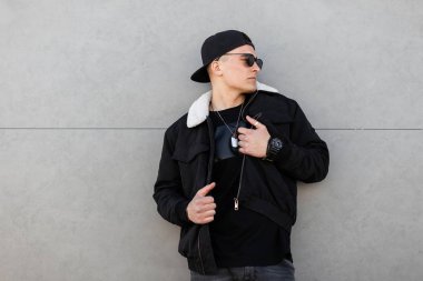 Kot pantolon beyaz kürk ile moda siyah ceket koyu moda güneş gözlüğü şık kap şık kap şık genç hipster adam bahar gününde şehirde gri bir duvar yakınında pozlar. Çekici modern adam.
