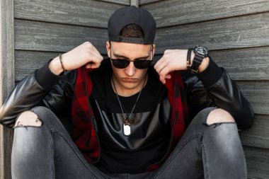 Siyah beyzbol şapkalı, siyah güneş gözlüklü, siyah tişörtlü yırtık kot pantolonlu, şehirde ahşap bir duvarın yanında duran yakışıklı hippi. Sokaktaki havalı adam. Sokak tarzı.