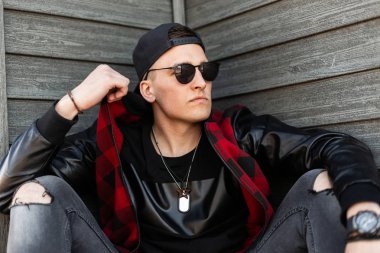 Siyah güneş gözlüğü ciddi yakışıklı genç hipster adam ahşap bir duvarın yakınında açık havada oturan bir kap gri yırtık kot siyah şık t-shirt bir moda damalı ceket içinde. İyi adam.
