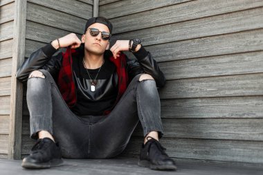 Siyah bir beysbol şapkası siyah bir hipster adam siyah bir t-shirt kırmızı damalı ceket bir vintage ahşap duvar yakınında oturan dinlenme. Havalı adam dışarıda. Sokak stili.