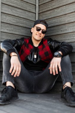 Spor ayakkabı içinde güneş gözlüğü gri şık kot moda kap moda kap Neşeli yakışıklı genç hipster adam oturur ve ahşap bir binanın yakınında bir yaz gününde gülümsüyor. Neşeli adam.