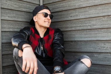 Koyu güneş gözlüğü yırtık gri kot şık bir damalı ceket şık bir beyzbol siyah kap yakışıklı neşeli genç hipster adam bir vintage ahşap binanın yakınında oturur. Çekici adam gülümseyen.