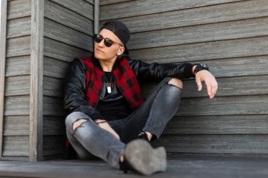 Moda yaylası şık siyah beyzbol şapkası şık giysiler içinde Modern Amerikan yakışıklı genç hipster adam oturur ve sıcak bir yaz günü sahiptir. Çekici erkek modeli rahatlatır. Erkek gençlik modası.