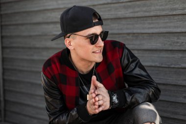 Şık güneş gözlüğü siyah bir kap bir vintage kırmızı damalı ceket Pozitif genç hipster adam oturur ve güneşli bir bahar gününde ahşap bir duvar yakınında gülümsüyor. Neşeli mutlu adam hafta sonu sahiptir.