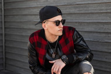 Gri yırtık kot bir trendy kırmızı kontrol ceket siyah güneş gözlüğü bir beyzbol şapkası sevimli genç hipster adam bir yaz günü ahşap bir binanın yakınında sokakta oturur. Amerikalı adam dinleniyor.