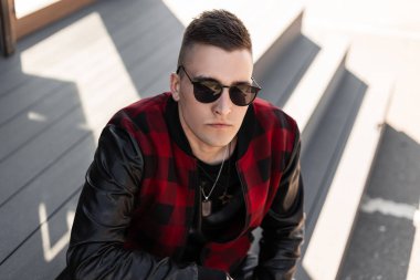 Siyah bir t-shirt kırmızı ve siyah vintage ceket koyu güneş gözlüğü şık bir saç modeli ile Cool genç adam hipster güneşli bir günde şehirde ahşap adımlar üzerinde oturur. İyi adam hafta sonu zevk.