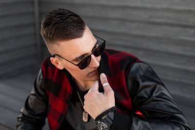 Ahşap bir vintage duvar yakınında poz şık güneş gözlüğü ile siyah bir t-shirt kırmızı trendy ekose ceket ile saç modeli ile Güzel genç hipster adam. Yakışıklı modern adam model.