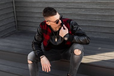 Siyah güneş gözlüğü ile trendy siyah t-shirt yırtık kot şık ekose ceket Yakışıklı genç hipster adam güneşli bir günde ahşap bir vintage duvar yanında oturur. Adam dinleniyor. Amerikan sokak stili