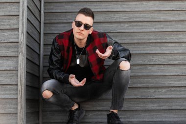 Modaya uygun bir ahşap binanın yakınında oturan poz moda spor ayakkabı yırtık şık kot siyah bir t-shirt moda yakışan bir moda ceket koyu güneş gözlüğü serin Amerikan genç adam. Çekici ciddi adam.