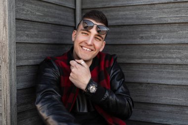Şık güneş gözlüğü bir trendy kırmızı ve siyah damalı ceket olumlu bir gülümseme ile Joyful yakışıklı genç hipster adam eski ahşap duvar yakınında oturuyor ve hafta sonu zevk. Neşeli adam.