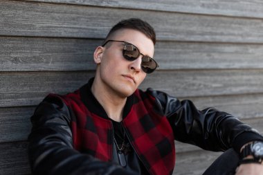 Vintage duvarda deri kollu şık kırmızı damalı ceket şık bir kırmızı damalı ceket şık koyu güneş gözlüğü ile ciddi bir çekici hipster genç adam Portresi. Yakışıklı adam dinleniyor