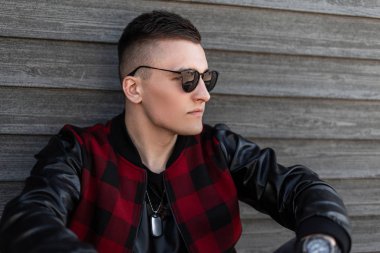 Ahşap bir duvara yakın deri kollu şık bir kırmızı damalı ceket şık bir saç modeli ile modern yakışıklı genç adam hipster trendy güneş gözlüğü Portresi. Çekici adam dinlenme.