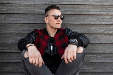Gri yırtık kot bir trendy ekose ceket şık bir saç modeli ile koyu güneş gözlüğü güzel genç hipster adam bir yaz gününde ahşap bir binanın yakınında açık havada oturur. Cool Amerikan adam dinleniyor.