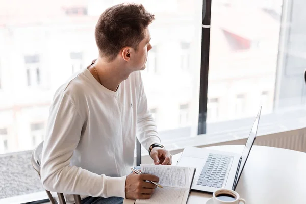 Pohledný mladý blogger s notebookem v bílé košili sedí u stolu v kanceláři a bere si poznámky do zápisníku. Pracujte na tvůrčím projektu. Vzdálená práce. — Stock fotografie