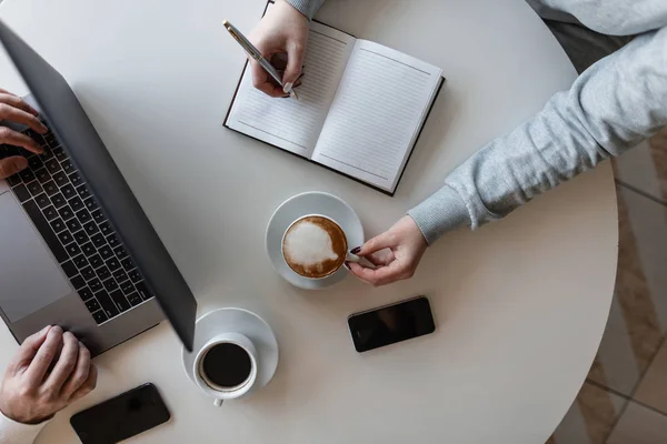 Νεαρή γυναίκα ελεύθερη επαγγελματίας κάθεται σε ένα τραπέζι με έναν επιχειρηματία σε ένα καφέ πίνοντας καφέ και κάνοντας εγγραφές σε ένα σημειωματάριο. Ομαδική δουλειά. Άποψη από ψηλά. — Φωτογραφία Αρχείου