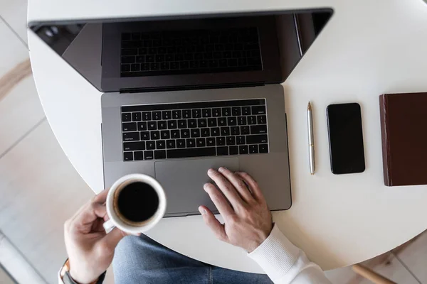 Obchodní muž pracující v laptopu a pije kávu, když sedí u stolu v kavárně. Chlápek pracuje vzdáleně u počítače. Pohled shora na plochu. — Stock fotografie