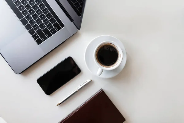 Kovový laptop, smartphone, zápisník, pero a šálek černé kávy na bílém dřevěném stole. Pohled shora na plochu. Moderní technologie. Vzdálená práce. — Stock fotografie