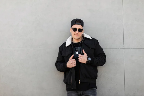 Joyful young hipster in a black baseball cap with sunglasses in a stylish black jacket with white fur in gray jeans is standing and smiling near the gray wall. Positive cute guy enjoying the weekend. — Stock Photo, Image