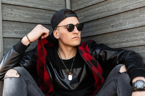 Hombre hipster joven guapo serio en gafas de sol negras en una chaqueta a cuadros de moda en una camiseta elegante negro en jeans rasgados grises en una gorra que descansa sentado al aire libre cerca de una pared de madera. Buen tipo. . — Foto de Stock