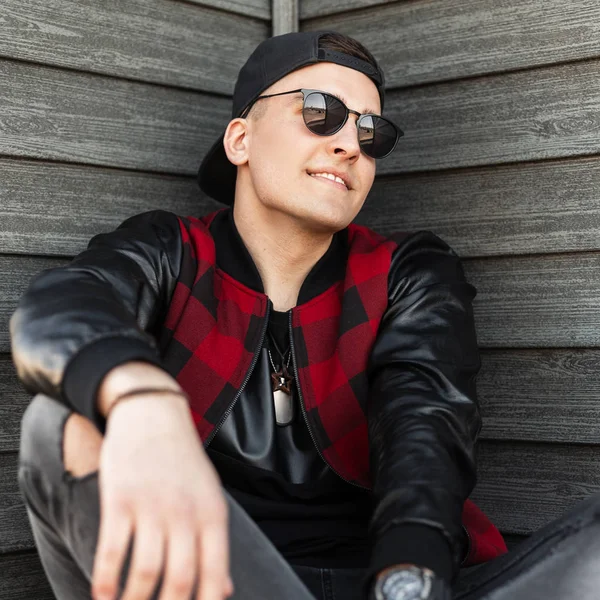 Joyful handsome young hipster man in torn gray jeans in a black cap in a trendy red and black checkered jacket in stylish sunglasses sits near a vintage wooden wall and enjoys the weekend. Happy guy. — Stock Photo, Image