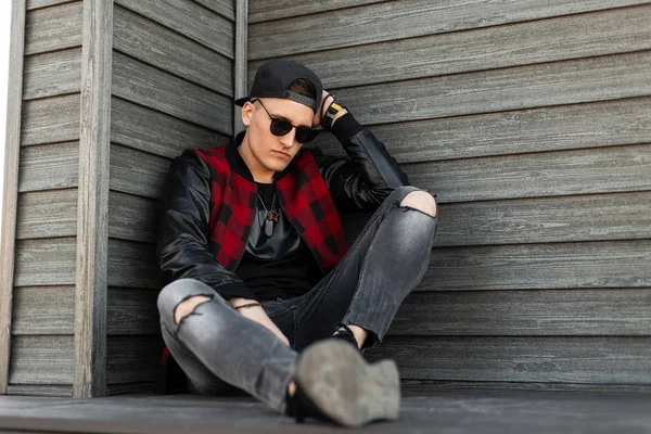 Atractivo joven hipster en gafas de sol oscuras de moda en una elegante chaqueta a cuadros roja y negra con mangas de cuero en jeans en zapatillas de deporte en una gorra se sienta cerca de una pared de madera vintage. Chico fresco descansando —  Fotos de Stock