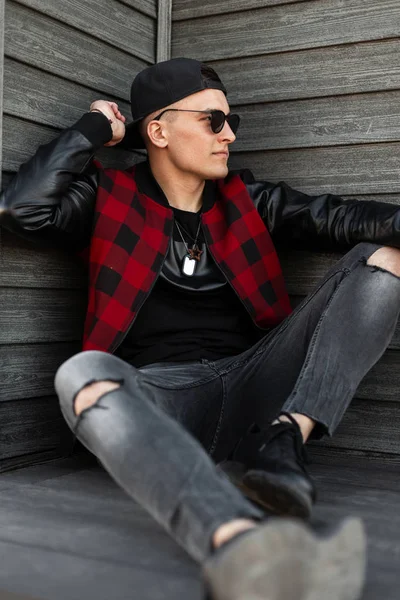 Elegante hombre hipster joven guapo en gafas de sol negras en una chaqueta a cuadros de moda en zapatillas de deporte en jeans rasgados grises en una gorra relajarse sentado al aire libre cerca de una pared de madera. Atractivo chico descansando . — Foto de Stock