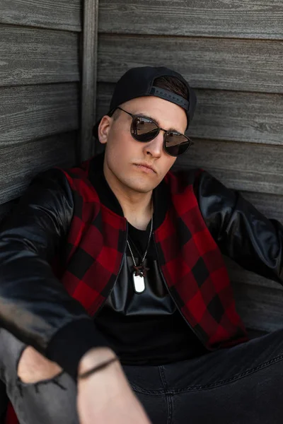 Joven hombre hipster guapo americano en ropa de moda en una elegante gorra de béisbol negro en gafas de sol negras se sienta cerca de una pared de madera y disfruta de un cálido día de verano. chico urbano es relajante . — Foto de Stock