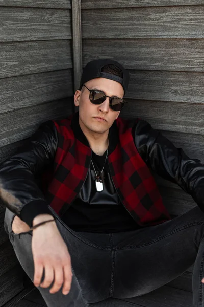 Guapo joven hipster hombre en una gorra negra en gafas de sol de moda en una chaqueta a cuadros rojo vintage en jeans rasgados grises se relaja sentado cerca de un edificio de madera. El tipo genial está descansando al aire libre. Estilo de calle . — Foto de Stock