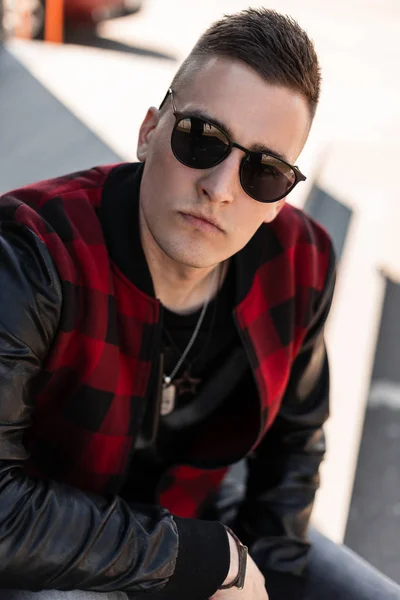 Retrato de un joven hipster serio con gafas de sol elegantes con un peinado de moda en una elegante chaqueta a cuadros roja con mangas de cuero al aire libre. Tipo atractivo descansando en un día soleado . — Foto de Stock