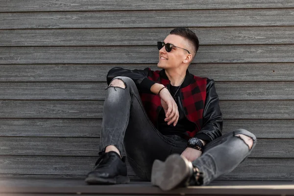 Happy American Young hipster man in een modieuze geruite jas met lederen mouwen in geripte stijlvolle jeans in Sneakers zit en geniet van het weekend in de buurt van een houten muur. Knappe kerel glimlachend. — Stockfoto