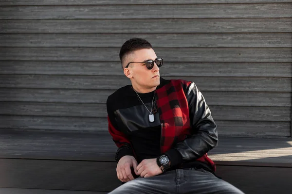 Hombre hipster guapo de moda en gafas de sol elegantes con una gorra de béisbol se sienta cerca de un edificio de madera en un día oscuro de primavera. Americano chico modelo es descansando al aire libre . — Foto de Stock