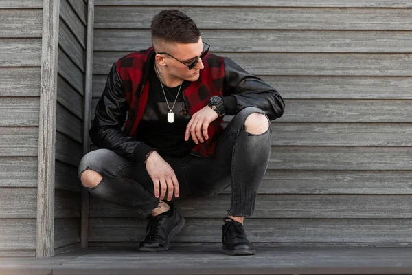 Der hübsche junge Hipster-Mann mit schwarzer Sonnenbrille, trendig rot karierter Jacke und Turnschuhen in stylischen zerrissenen Jeans sitzt draußen an einer Holzwand. Amerikaner ruht sich aus. — Stockfoto