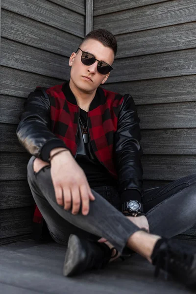 Stylischer junger Hipster-Mann mit schwarzer Sonnenbrille, modisch karierter Jacke und Turnschuhen in grauer zerrissener Jeans sitzt draußen an einer alten Holzwand. Amerikaner ruht sich gern aus. — Stockfoto