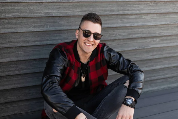 Joven hipster guapo alegre en vaqueros grises rasgados de moda en una chaqueta a cuadros rojo y negro de moda en gafas de sol elegantes se sienta cerca de una pared de madera vintage y disfruta del fin de semana. Tipo positivo . — Foto de Stock