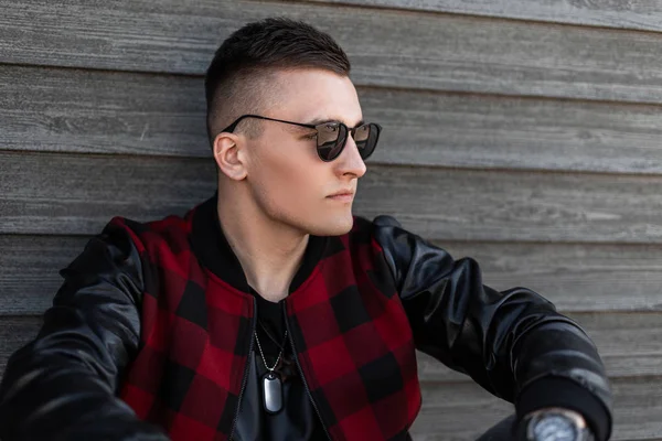 Retrato de un joven guapo moderno hipster gafas de sol de moda con un peinado de moda en una elegante chaqueta a cuadros roja con mangas de cuero cerca de una pared de madera. Atractivo chico descansando . — Foto de Stock