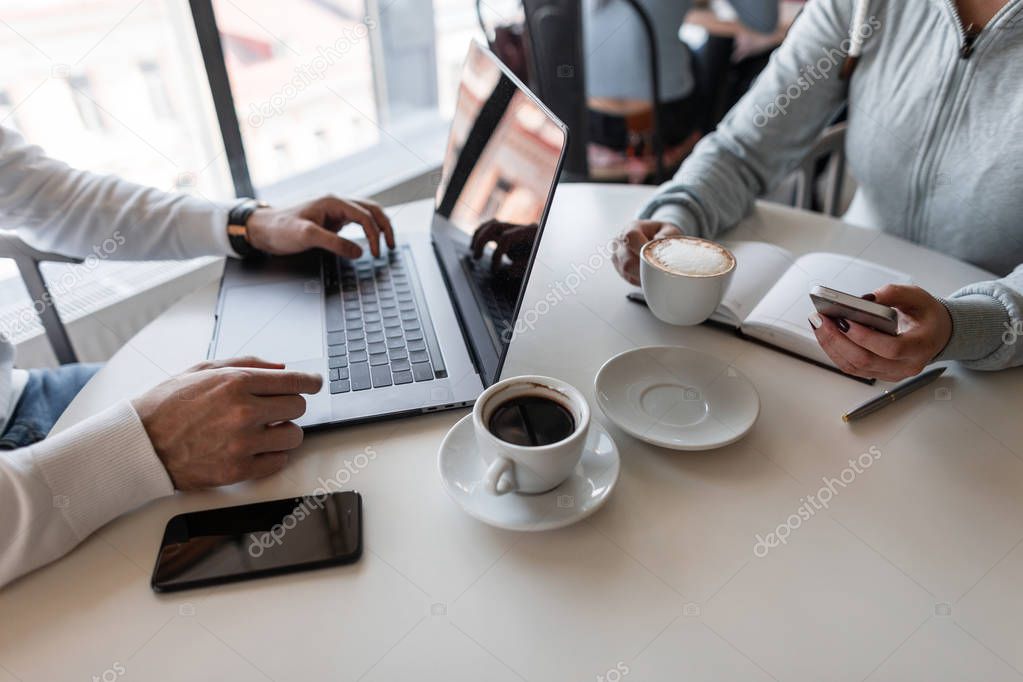 Two successful freelancers sitting in cafes working on a creative project for a cup of coffee. Closeup of male hands with a laptop and female hands with a phone and a cup of coffee. Remote work.