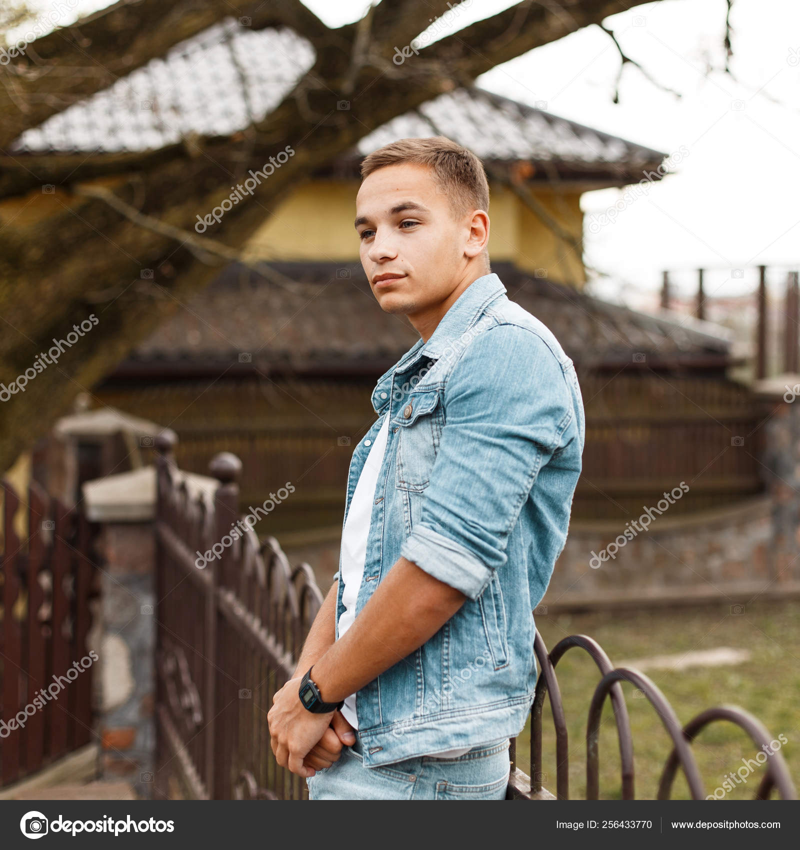 Flot ung med blå øjne i fashionabel denim jakke i hvid t-shirt i trendy jeans nær et vintage nær et træhus. Attraktiv fyr model udendørs . — Stock-foto ©