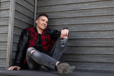 Mutlu Amerikan şık genç adam bir vintage ahşap binanın yakınında oturan rahat spor ayakkabı yırtık şık kot deri kollu şık bir damalı ceket. Neşeli adam modeli.