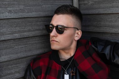 Ahşap bir duvara yakın deri kollu şık bir kırmızı ve siyah damalı ceket şık bir saç modeli ile şık siyah gözlük çekici genç hipster adam. Bir adamın yüzünün portresi. 