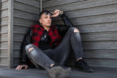 Siyah spor ayakkabı gri yırtık kot bir trendy kırmızı damalı ceket güneş gözlüğü ile şık bir saç modeli ile Ciddi genç hipster adam ahşap bir binanın yakınında oturan rahatlatır. İyi adam dinleniyor..