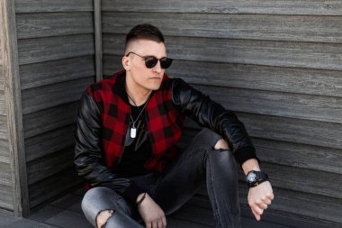 Gri yırtık kot bir moda kırmızı ekose ceket şık bir saç modeli ile siyah güneş gözlüğü güzel genç hipster adam bir yaz gününde ahşap bir binanın yakınında açık havada oturur. Amerikalı adam dinleniyor.