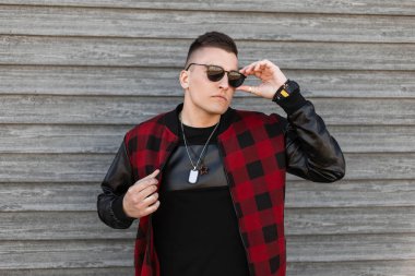 Şık bir genç Amerikan hipster adam bir bahar gününde ahşap bir vintage binanın yakınında poz tılsımları ile şık güneş gözlüğü bir ekose ceket. Bir yürüyüş için yakışıklı çekici adam.