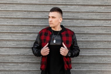 Modayı takip eden siyah ve kırmızı kareli ceketli, modaya uygun siyah tişörtlü ve muskalı çekici genç bir adam yaz günü eski bir ahşap duvarın yanında poz veriyor. Şık bir adam. Amerikan sokak tarzı erkek giyim